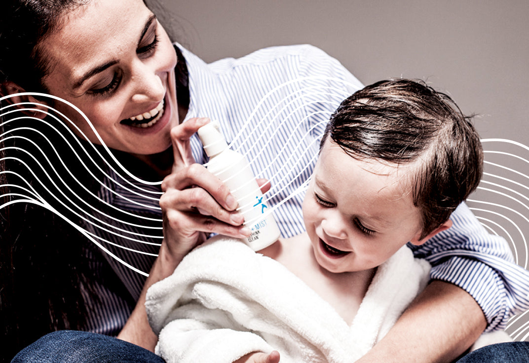Woman holding a little kid wrapped in a towel with a mist bottle in one hand pointed at the kid, both smiling.