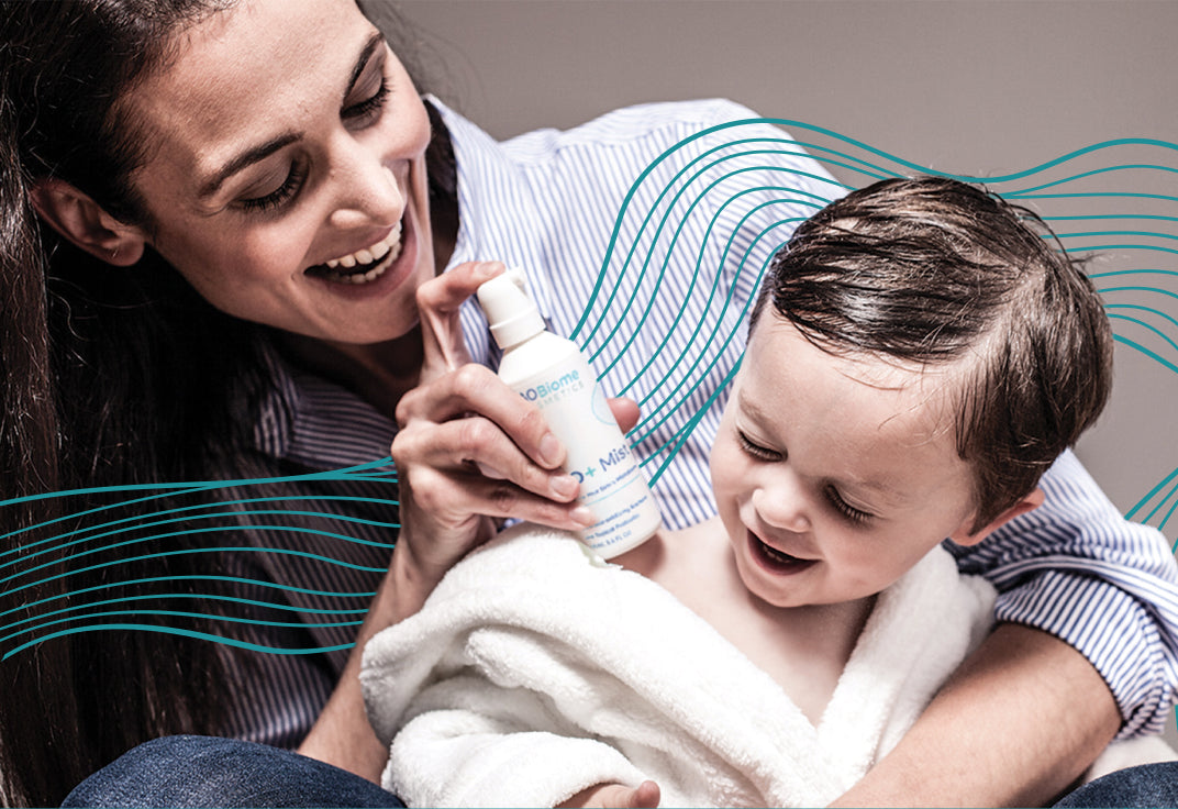 Woman holding a little kid wrapped in a towel with a mist bottle in one hand pointed at the kid, both smiling.