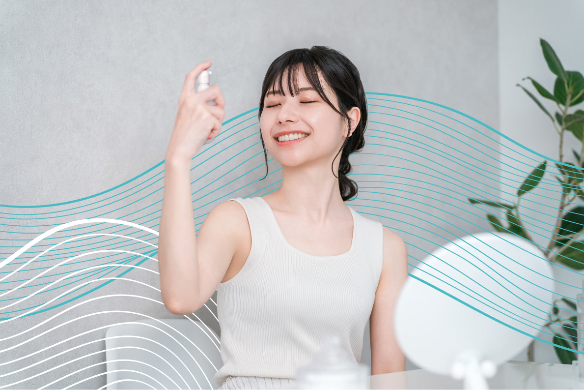 Woman using a spray bottle aimed at her face, smiling with her eyes closed, sitting at a table with a mirror on it.