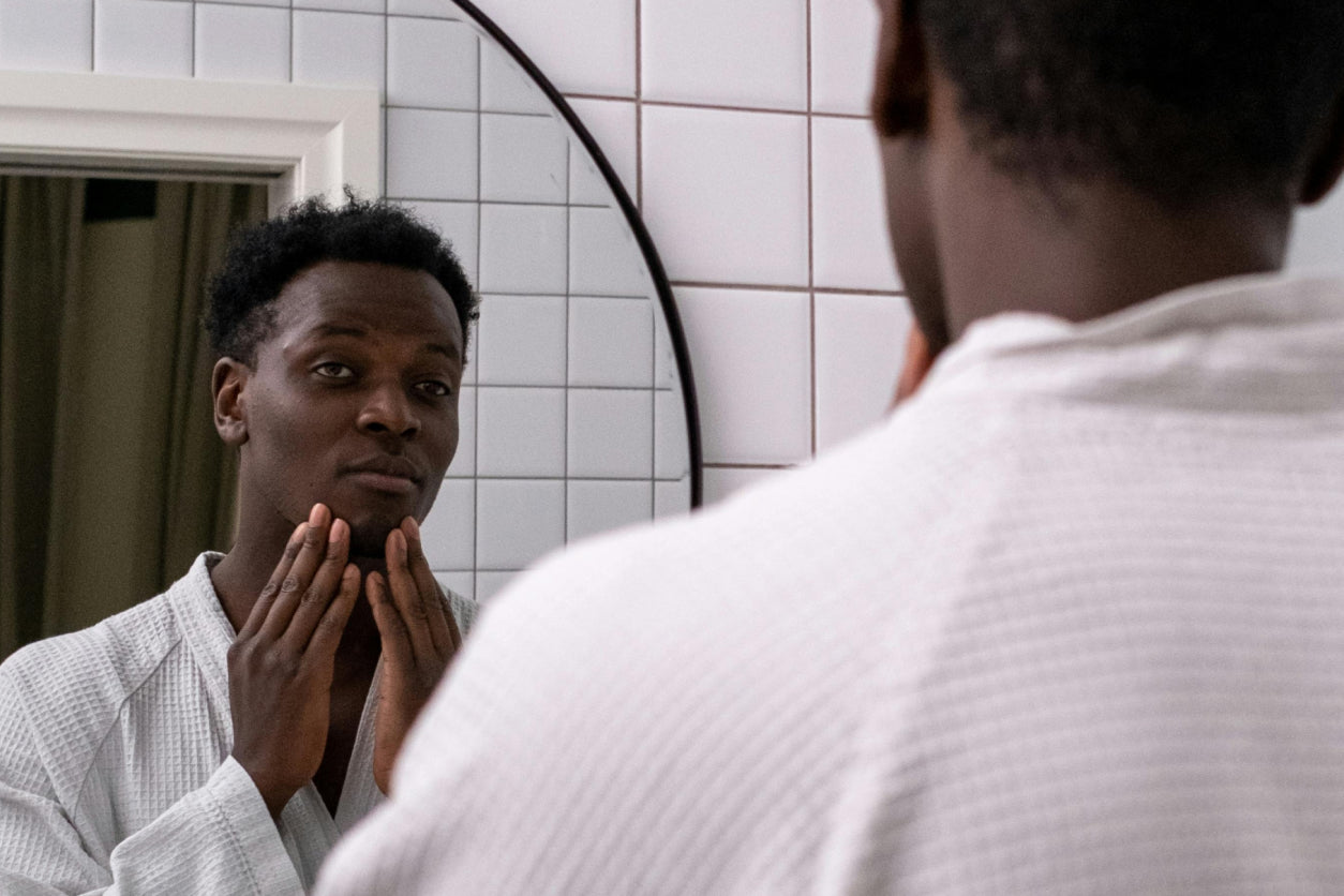 Man in a bathrobe with both hands on his chin, examining his face in a bathroom mirror.