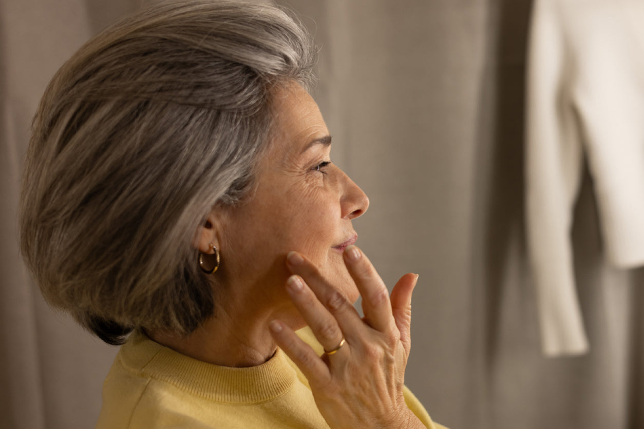 Woman with gray hair wearing a yellow sweater, looking to the side while touching her above her jawline with her finger.