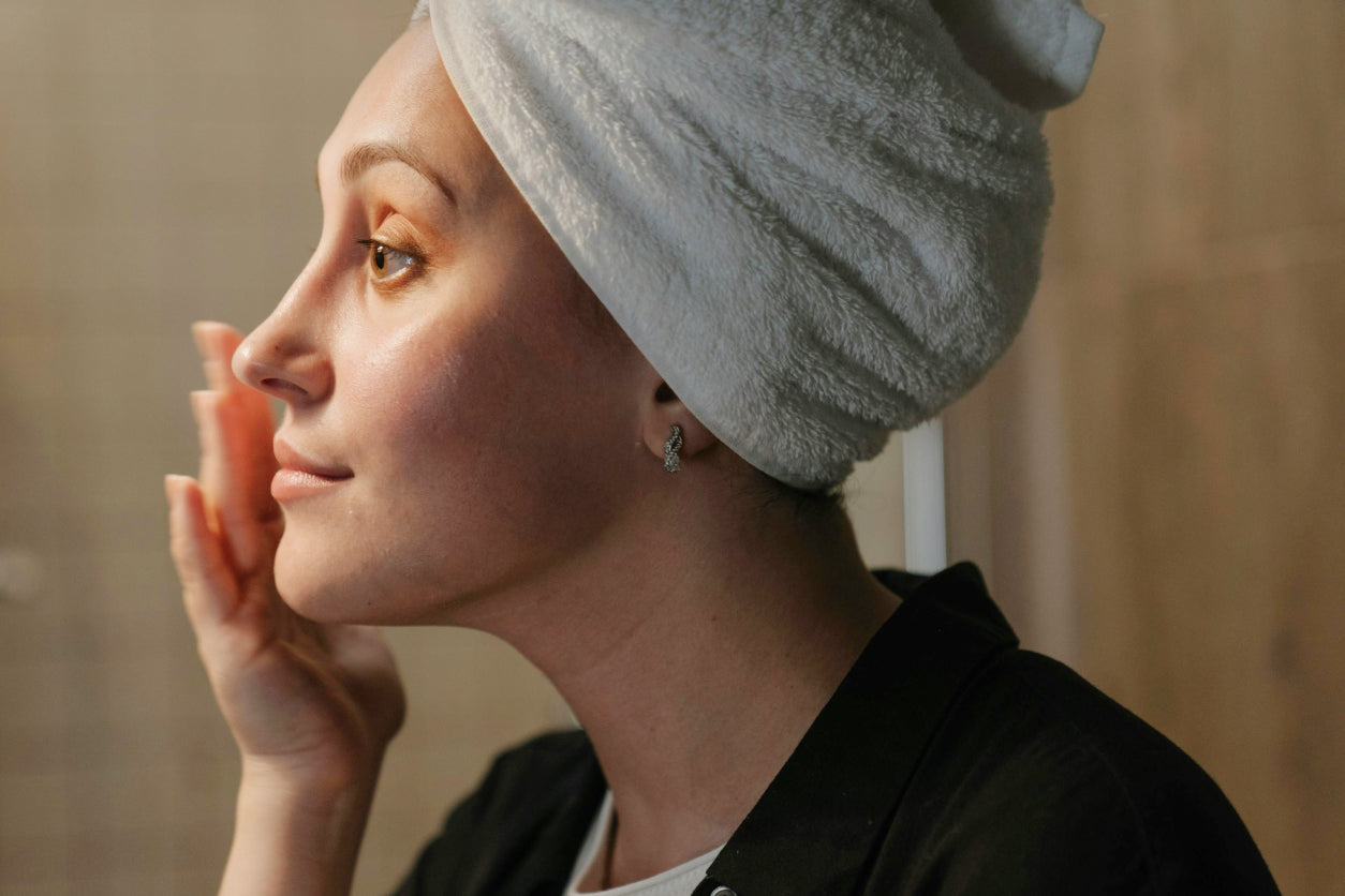 Woman with a towel wrapped around her hair, patting one side of her face with her hand.